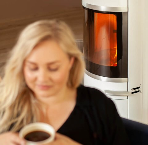 Blonde woman sitting in front of a white wood-burning stove Scan 83-2 with a cup of coffee in her hands
