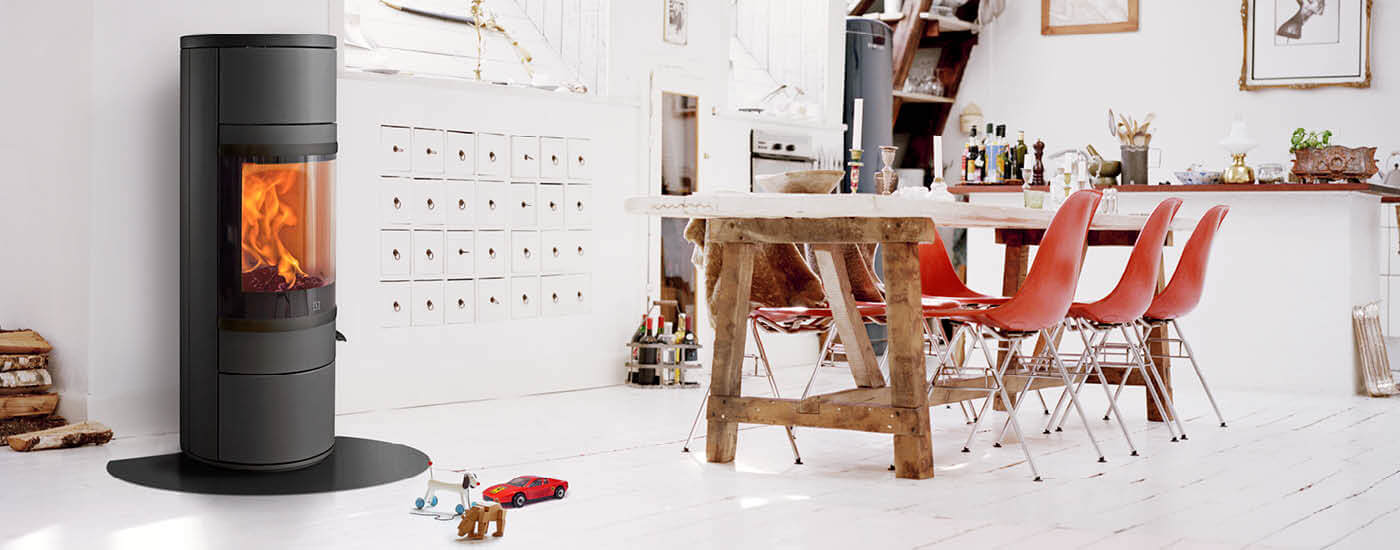 Zwarte ronde houtkachel in een witte keuken met een rustieke tafel en rode stoelen
