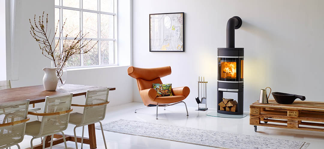 Zwarte ovale Scan 68 houtkachel in een witte kamer met een designstoel in helder leer en een ruwe pallettafel met wielen