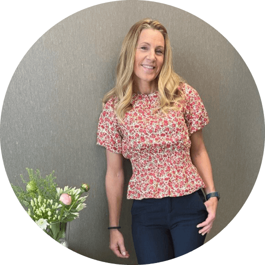 Blonde vrouw met donkere broek en bloemenblouse leunend tegen een muur, naast een glazen tafel met bloemen