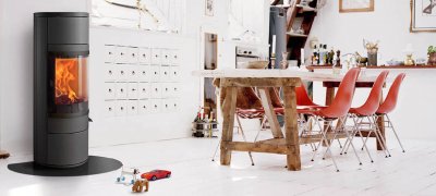 Zwarte ronde houtkachel in een witte keuken met een rustieke tafel en rode stoelen