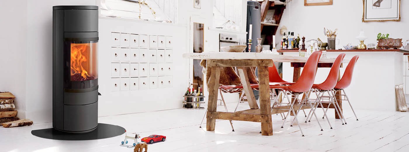 Zwarte ronde houtkachel in een witte keuken met een rustieke tafel en rode stoelen